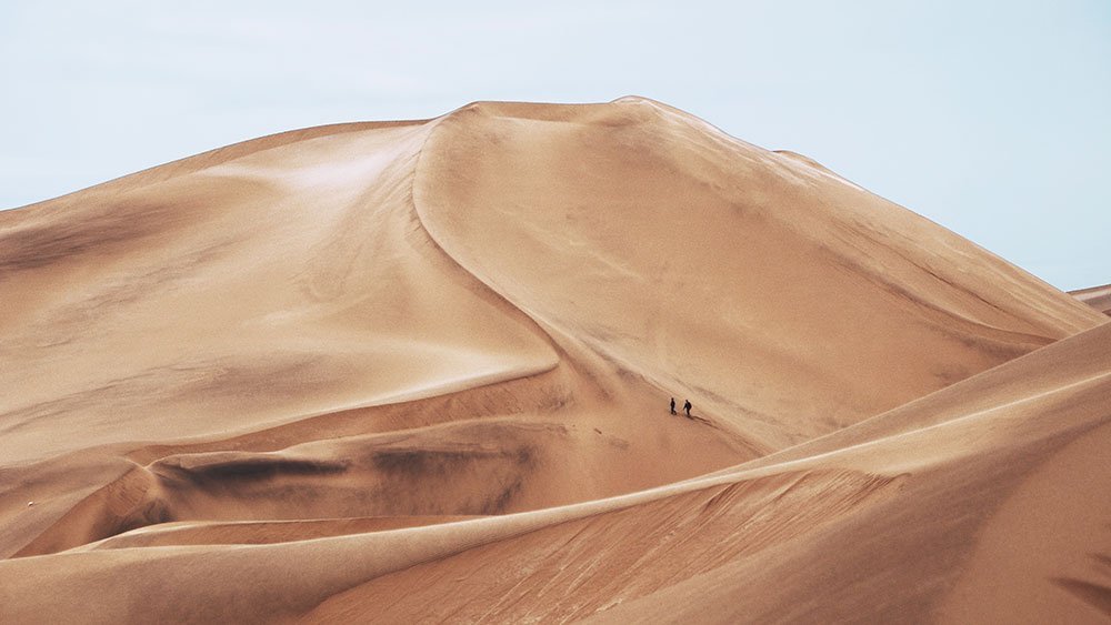 Two people hiking on a vast, curving sand dune in a desert landscape